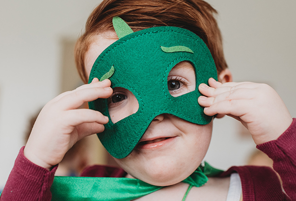 Ein kleiner Junge mit roten Haaren hält sich eine grüne Filzmaske vor das Gesicht und trägt passend dazu einen grünen Superheldenumhang.