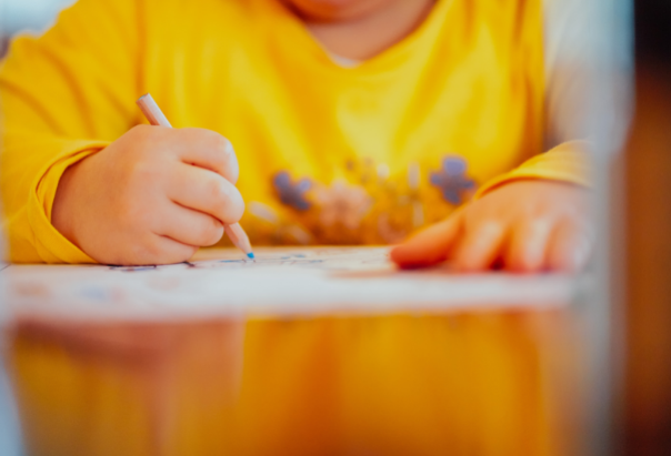 Ein Kind im gelben Sweatshort schreibt mit einem blauen Buntstift etwas zum Muttertag auf ein Blatt Papiier.