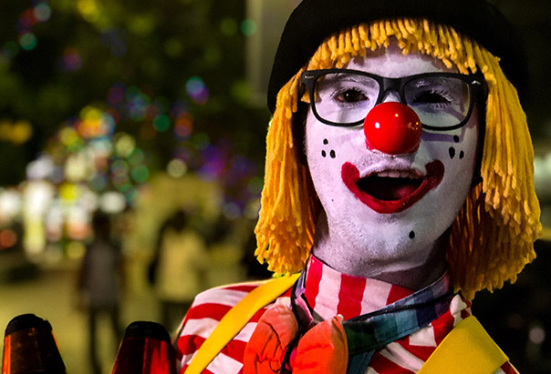 Ein Kind, das als Clown verkleidet ist, schaut in die Kamera und scheint dabei etwas zu sagen. Im Hintergrund erkennt man die verschwommenen Lichter einer einladenden nächtlichen Straße.
