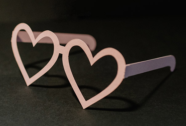 Auf einer dunklen Oberfläche liegt eine selbstgebastelte, rosafarbene Brille aus Papier in Herzform.