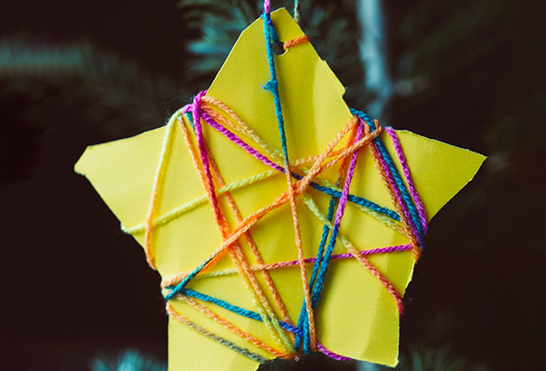 An einem Weihnachtsbaum hängt ein selbstgebastelter Sternanhänger aus gelbem Papier und buntem Wollfaden.