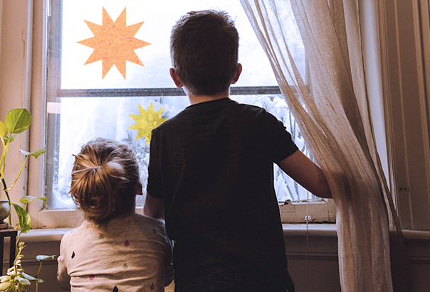 Zwei kleine Kinder stehen am Fenster und bewundern die Winterlandschaft. Auf der Fensterscheibe kleben bunte Sterne.