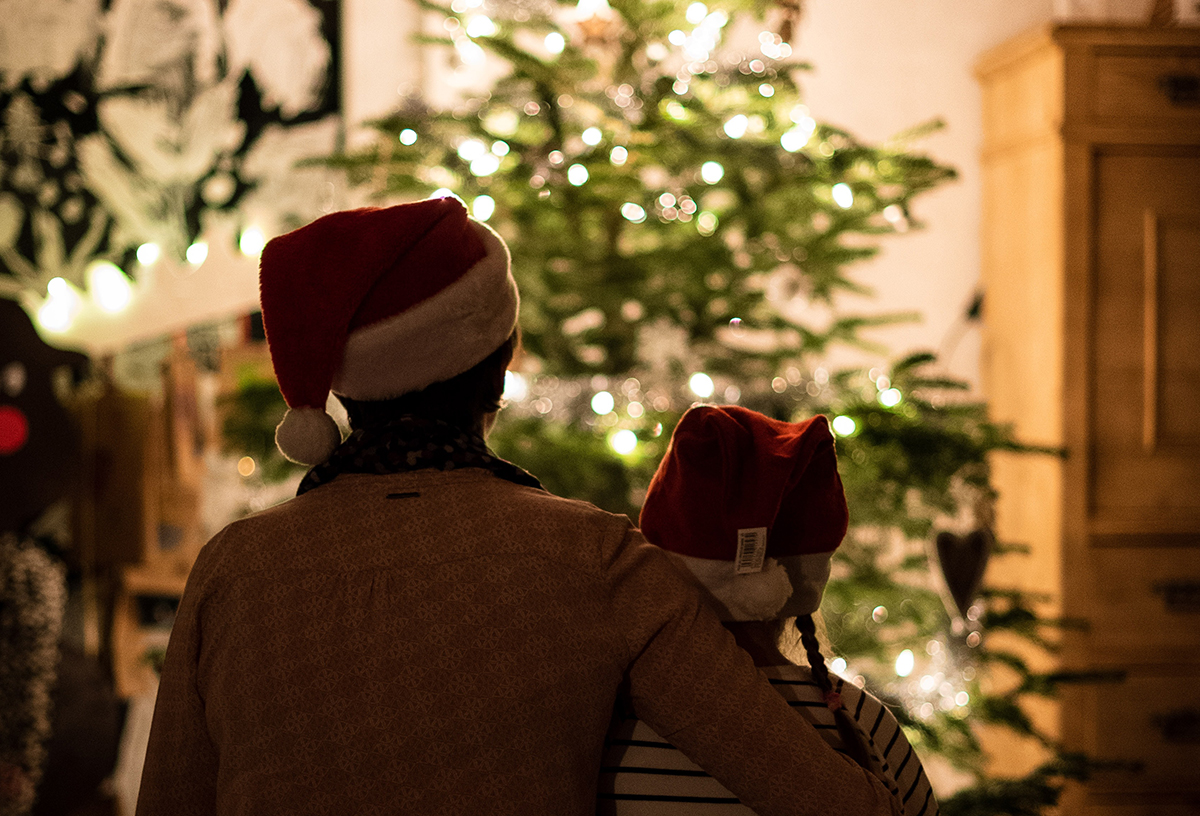 Eine junge Mutter sitzt mit ihrem Kind vor dem hell erleuchteten Weihnachtsbaum. Beide tragen Weihnachtsmützen.