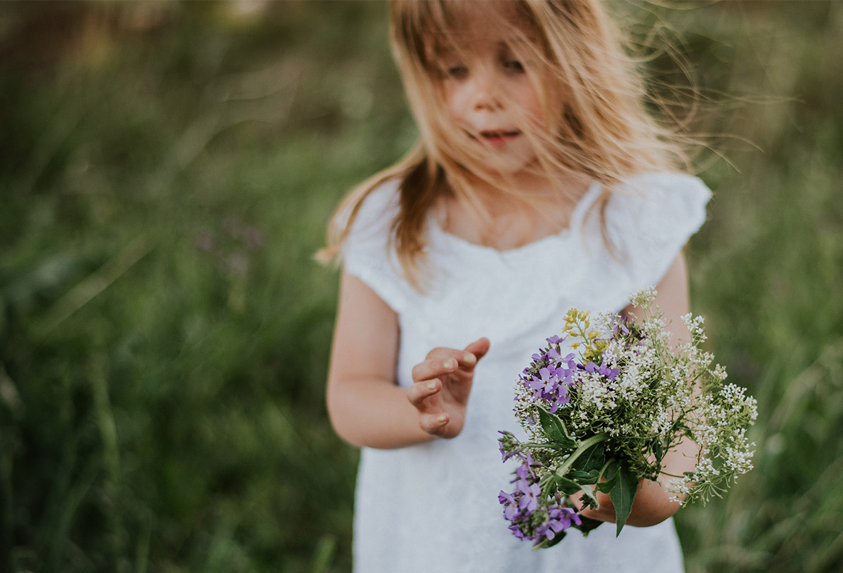 Ein kleines Mädchen in einem weißen Kleid steht auf einer Wiese und hält einen selbst gepflückten Blumenstrauß in der Hand.