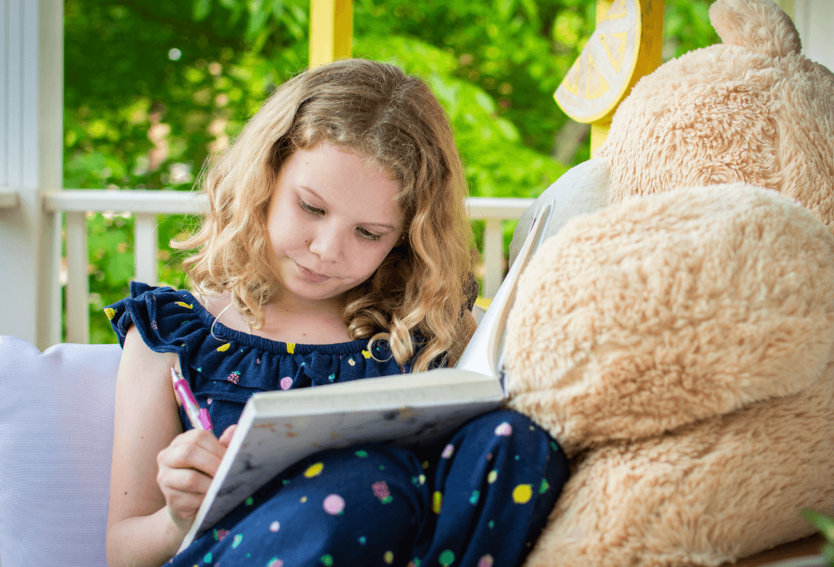 Ein kleines Mädchen sitzt mit ihrem Teddybären an einem sonnigen Tag draußen und schreibt in ihr Ferientagebuch.